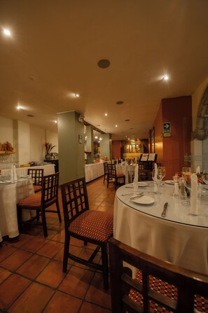 A restaurant with multiple dining tables, chairs, and a kitchen area in the background. - Hotel San Agustin El Dorado (Cusco)