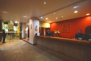 Zona de recepción de hotel con un mostrador de madera, puerta de cristal de entrada y pared roja con un logotipo.