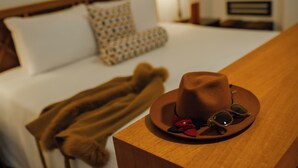 Una habitación de hotel con una cama bien hecha, un sombrero marrón y gafas de sol sobre una mesita de noche de madera.