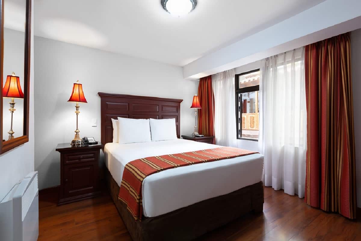 A hotel room with a large bed, wooden headboard, two bedside tables with lamps, a nightstand, and a window with curtains.