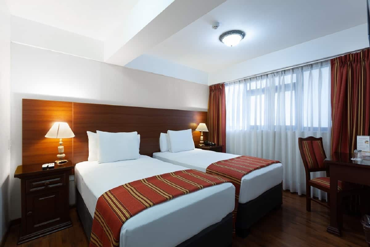 A hotel room with two beds, wooden headboards, a desk with a chair, and a window with curtains.
