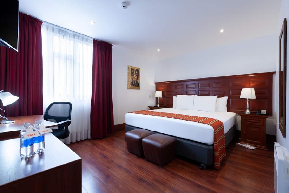 A hotel room with a large bed, a desk with a chair, a desk lamp, a TV, and a window with red curtains.