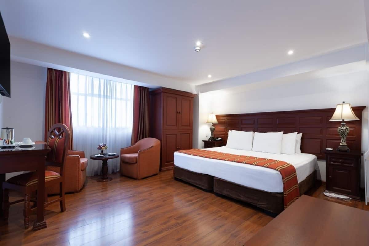 A hotel room with a bed, a desk, a chair, a small table, and a lamp.
