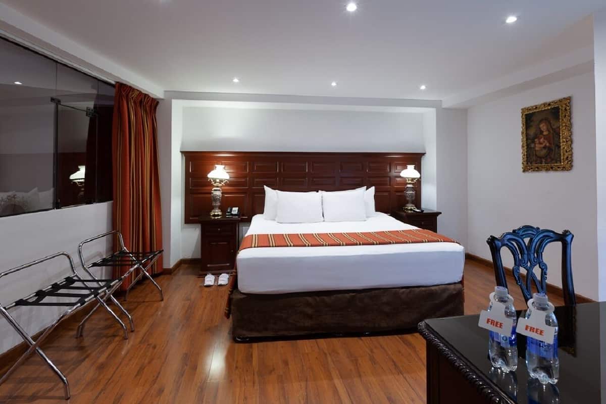 A hotel room with a large bed, wooden flooring, a desk with two bottles of water, and a chair.