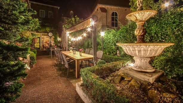 Un jardín al aire libre con mesa y sillas, una fuente decorativa y luces colgantes.