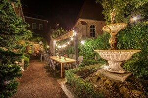Un jardín al aire libre con mesa y sillas, una fuente decorativa y luces colgantes.