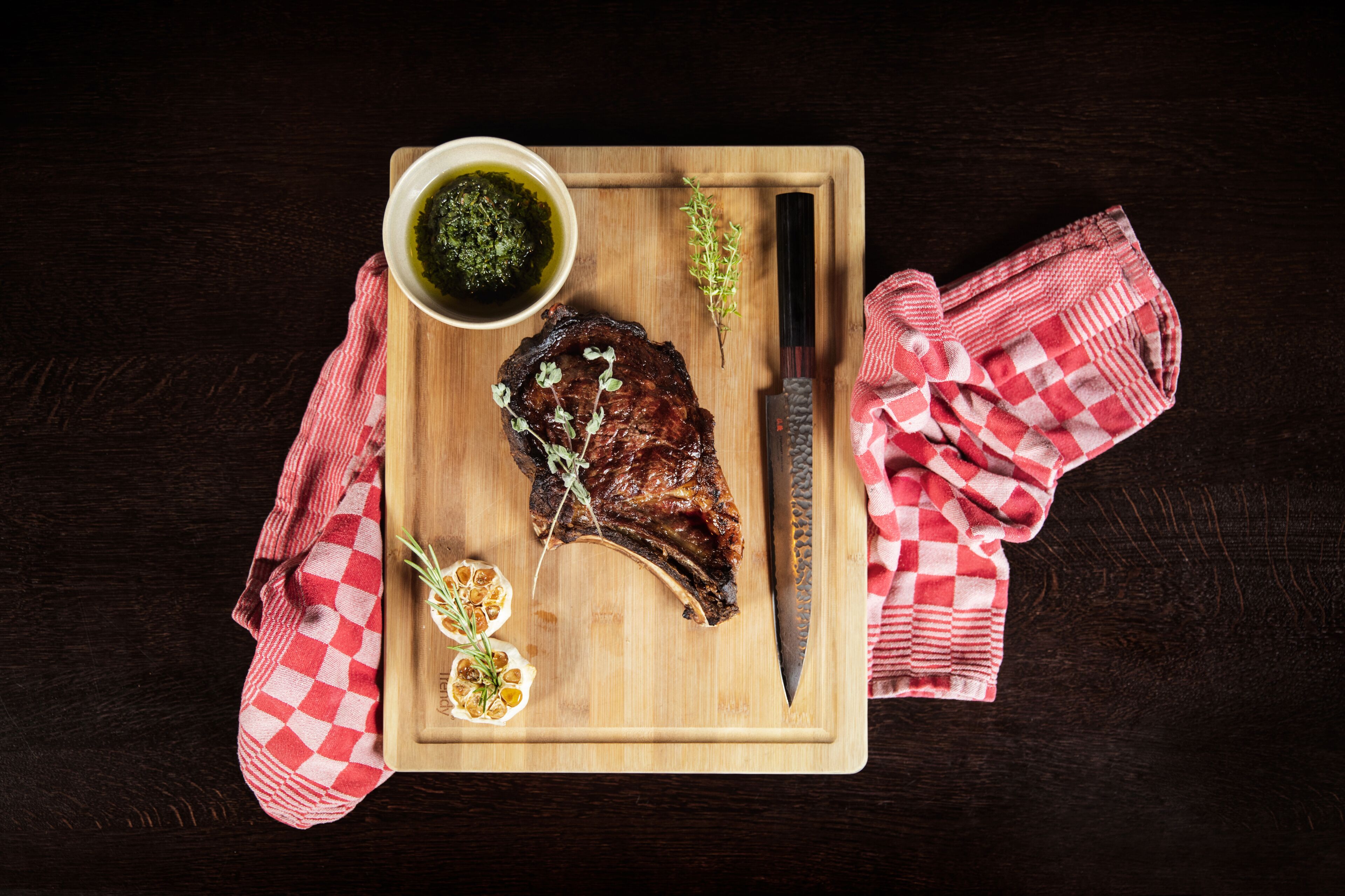 Un bistec sobre una tabla de madera, un cuchillo, un tazón de salsa y un mantel a cuadros rojos y blancos.