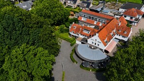 An aerial view of a residential area with a large building featuring a curved roof and a circular driveway. - Paping Hotel & Spa by Flow (Ommen)
