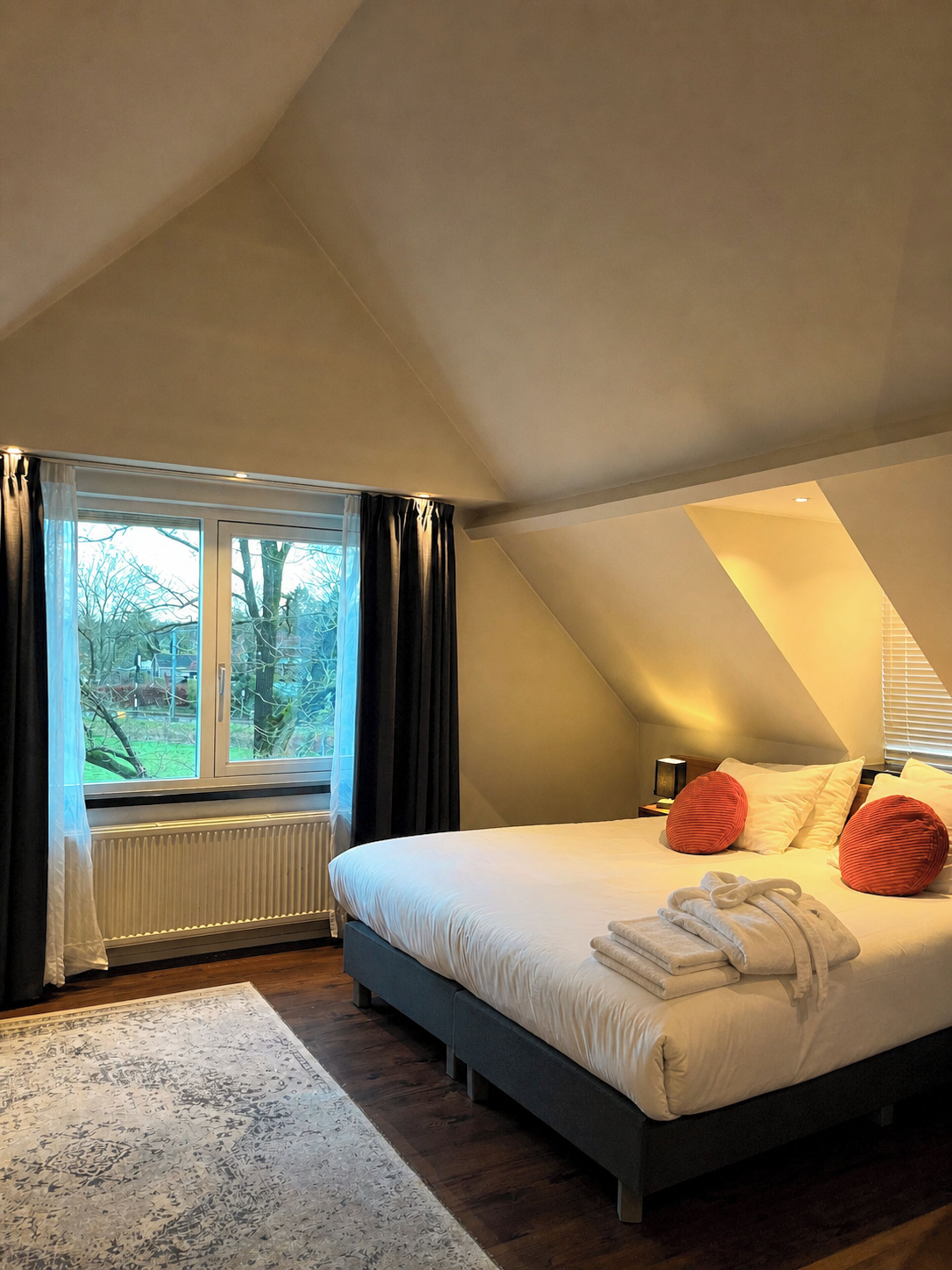 A bedroom with a large bed, a window with curtains, a radiator, and a wooden floor.