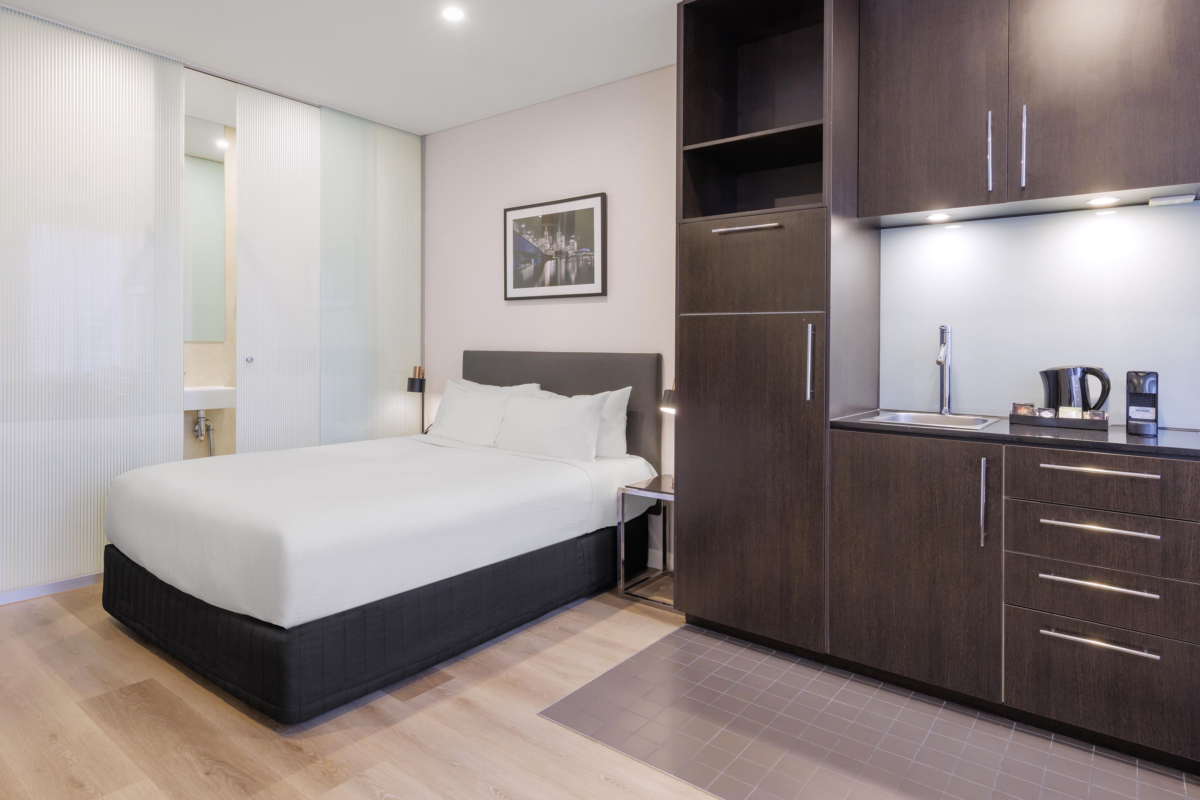 Moderni hotellihuone, jossa on sänky, yöpöytä, pieni sivupöytä ja pieni keittiöalue.