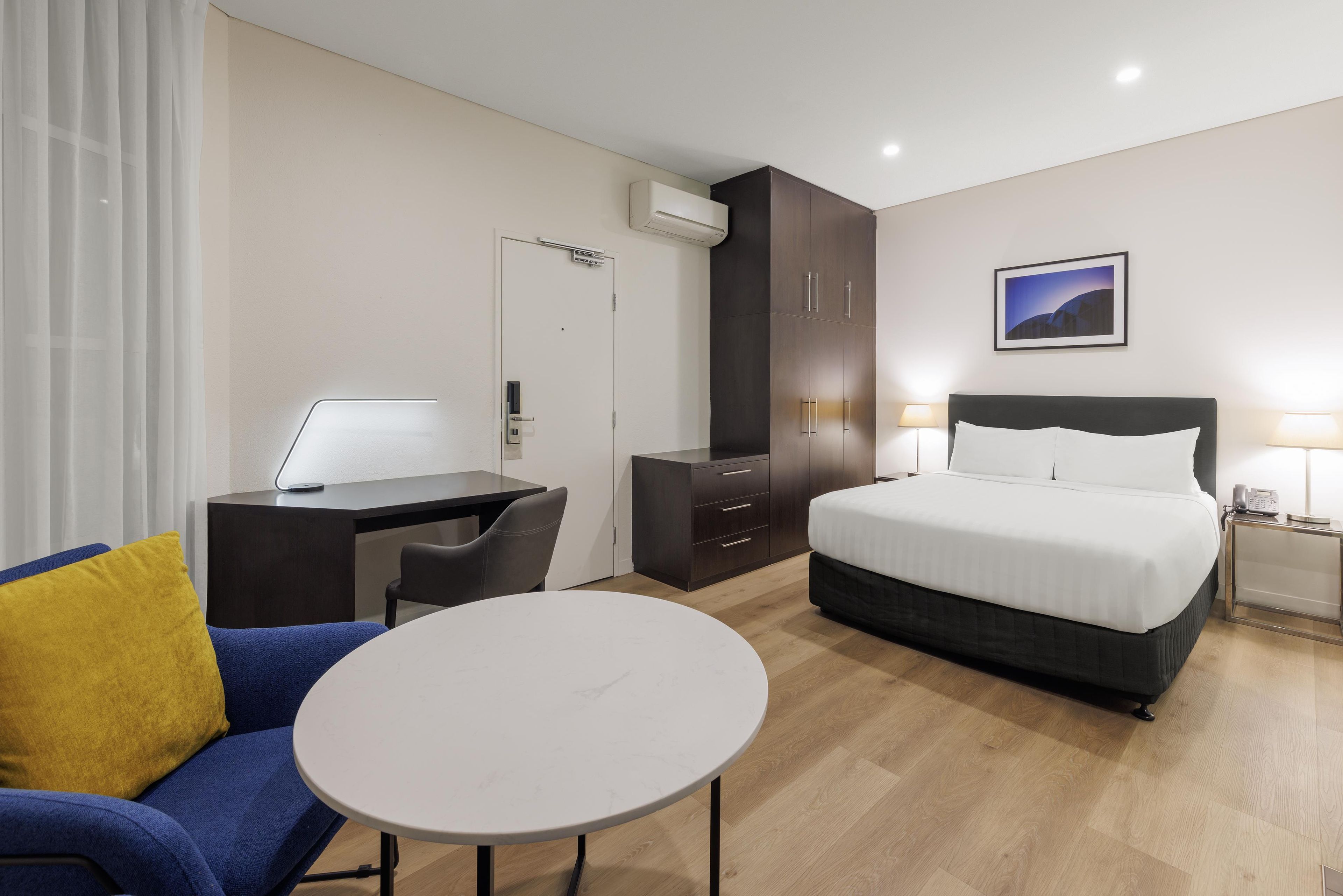 A hotel room with a bed, a desk, a chair, a small table, and a wardrobe.
