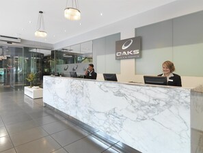 A hotel reception desk with marble countertops, a glass wall, and a sign for OAKS Hotels & Resorts. - Oaks Melbourne on Collins Suites (Melbourne)