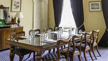 A dining room with a large wooden table, six chairs, a chandelier, and a window with curtains.