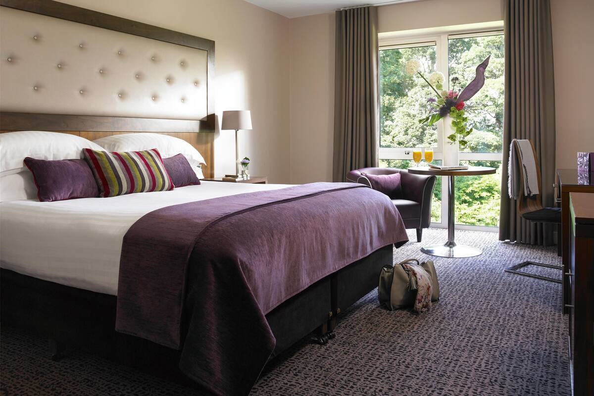 A hotel room with a large bed, a small table with a vase and two glasses, a chair, and a window with curtains.