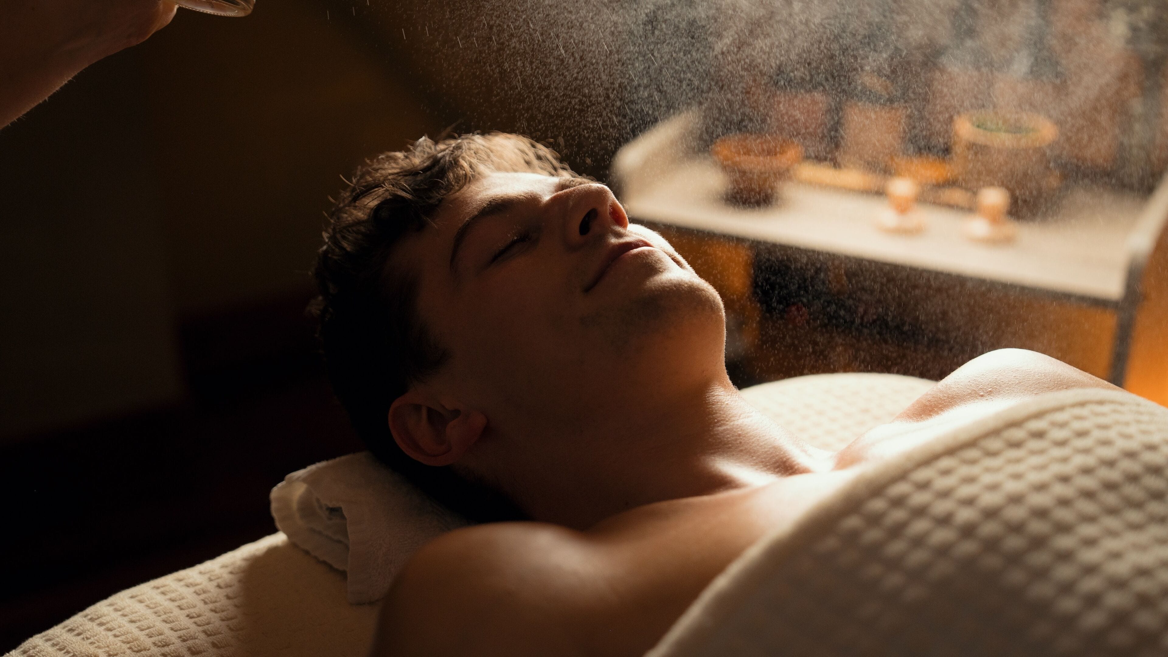 A person lying down, possibly in a spa or treatment room, with a table and some items in the background.