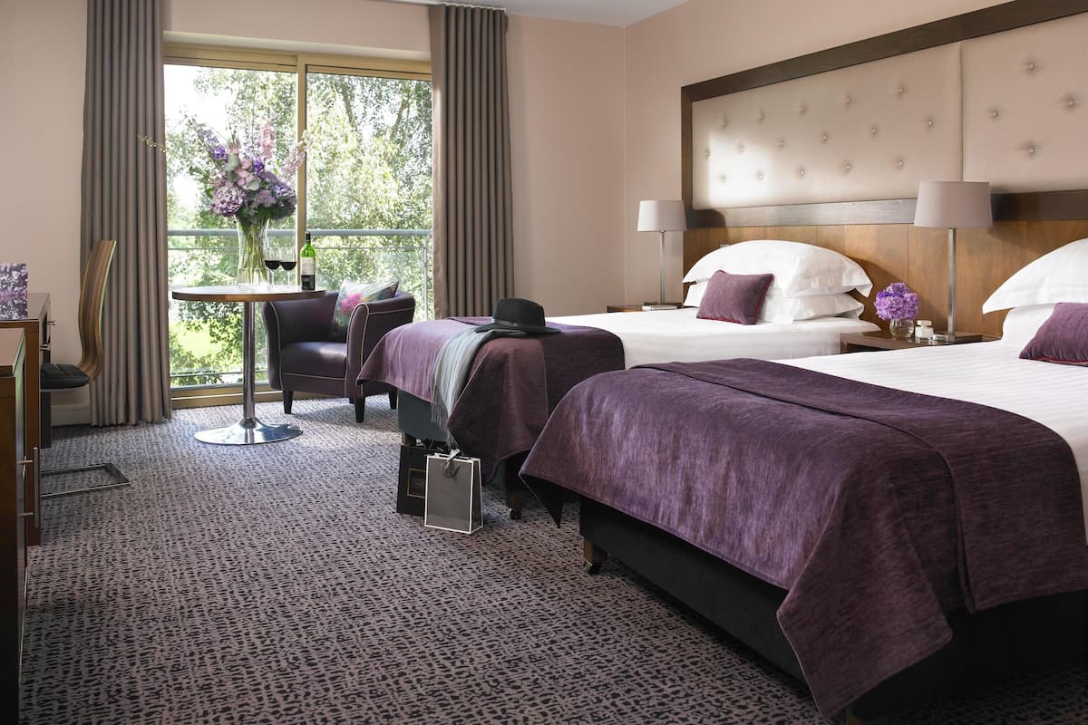 A hotel room with two beds, a sitting area with a table and chair, and a view of trees through the window.