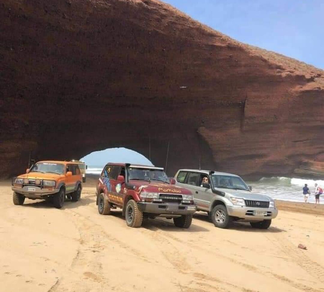 Drei Geländewagen parken auf einem sandigen Strand, im Hintergrund eine große Felsformation.