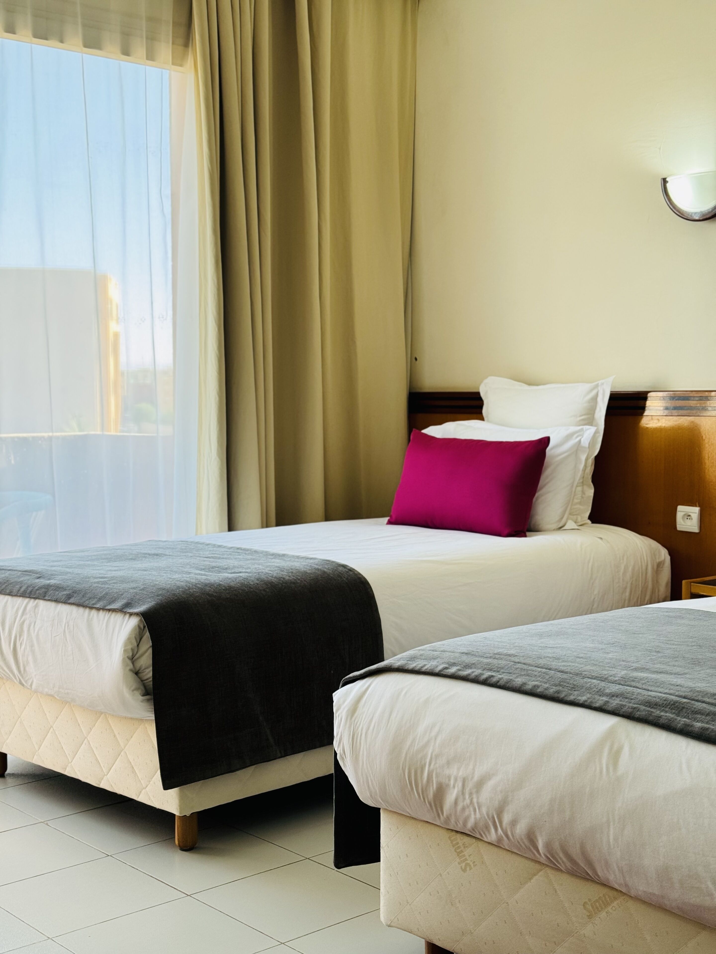 A hotel room with two beds, beige walls, a window with curtains, and a wooden headboard.