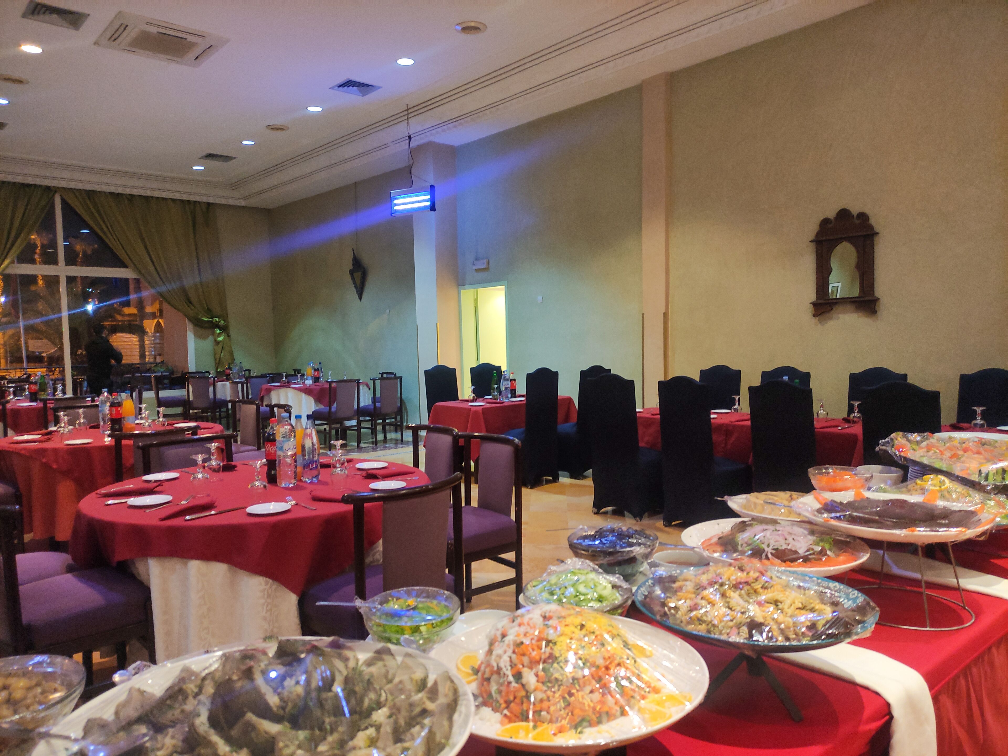 Une salle de banquet avec des tables rondes recouvertes de nappes rouges, des chaises habillées de housses noires et divers plats présentés sur des plateaux.