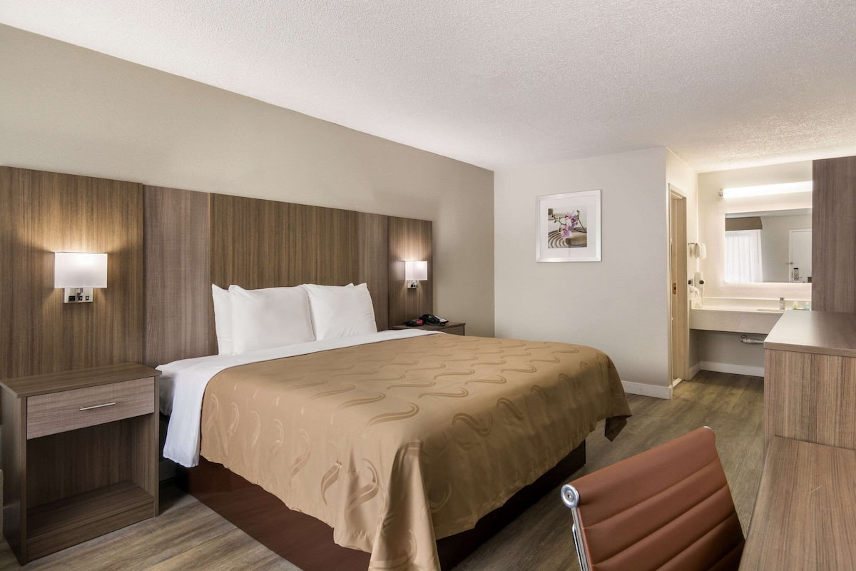 Une chambre d’hôtel avec un grand lit, une table de chevet, un bureau, une chaise et une salle de bain visible à travers une porte ouverte.