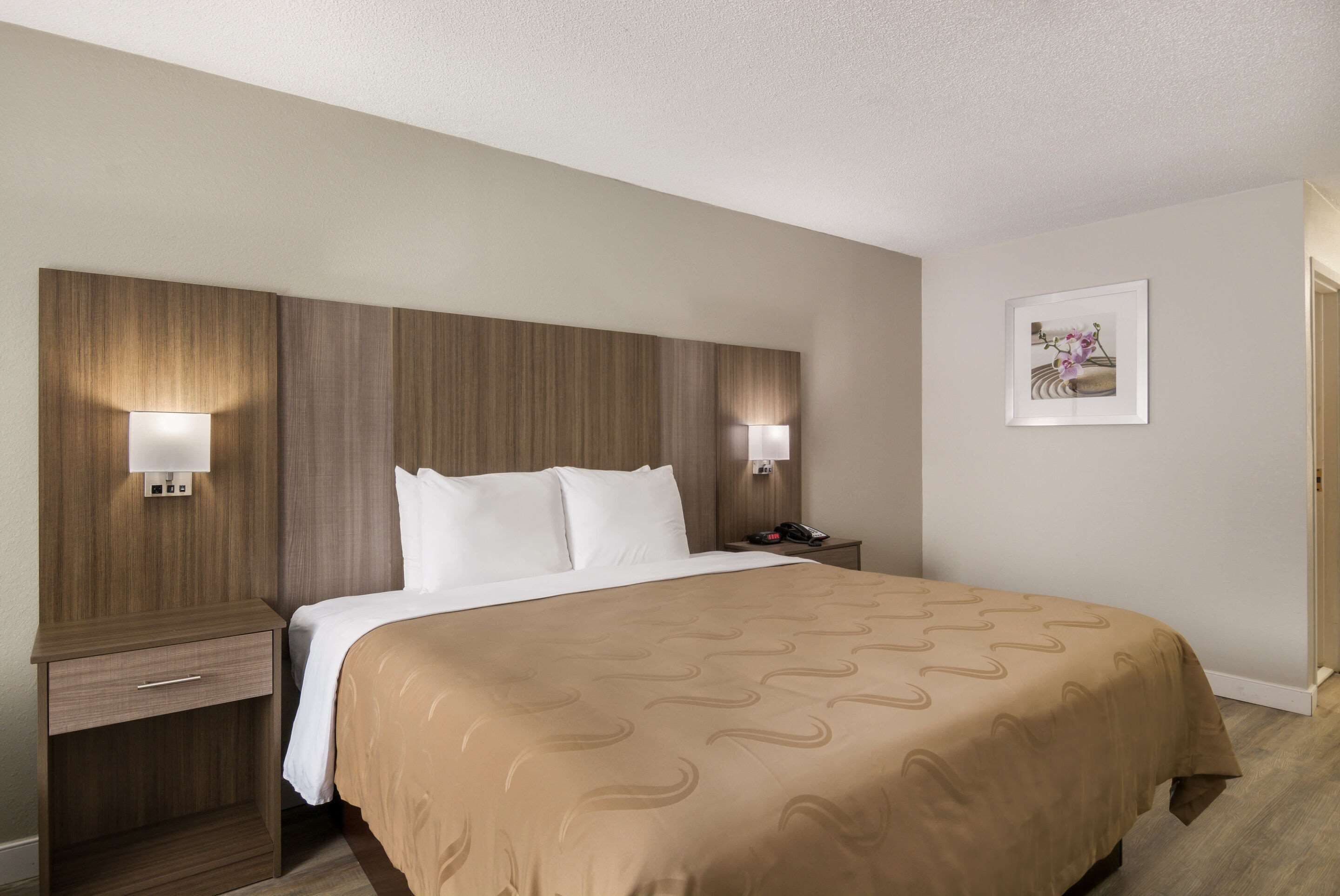 A hotel room with a large bed, two bedside tables, a nightstand, and a wall-mounted light fixture.