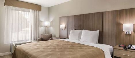 Quarto de hotel com uma cama grande, mesa de cabeceira com abajur, cadeira e janela com cortinas finas.
