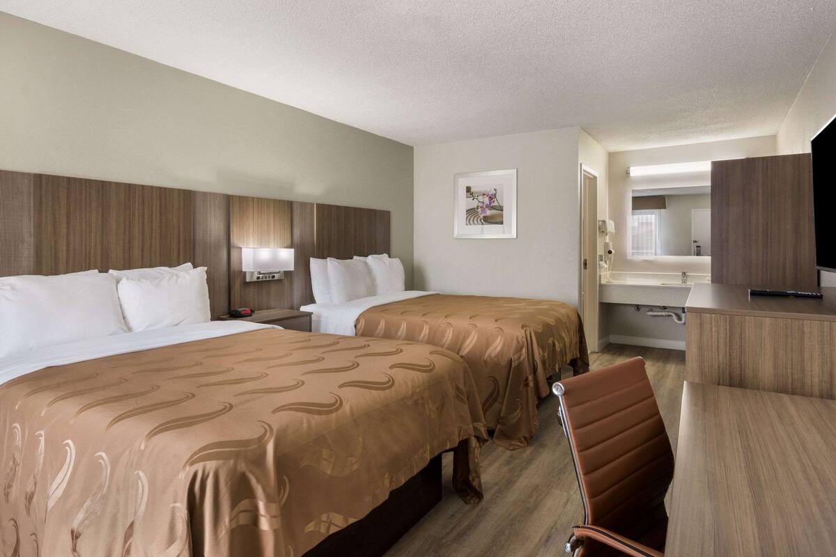 Une chambre d’hôtel avec deux lits, un bureau, une chaise, une télévision et une salle de bain visible à travers une porte ouverte.
