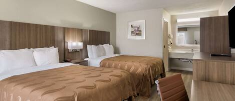 A hotel room with two beds, a desk, a chair, a TV, and a bathroom visible through an open door.