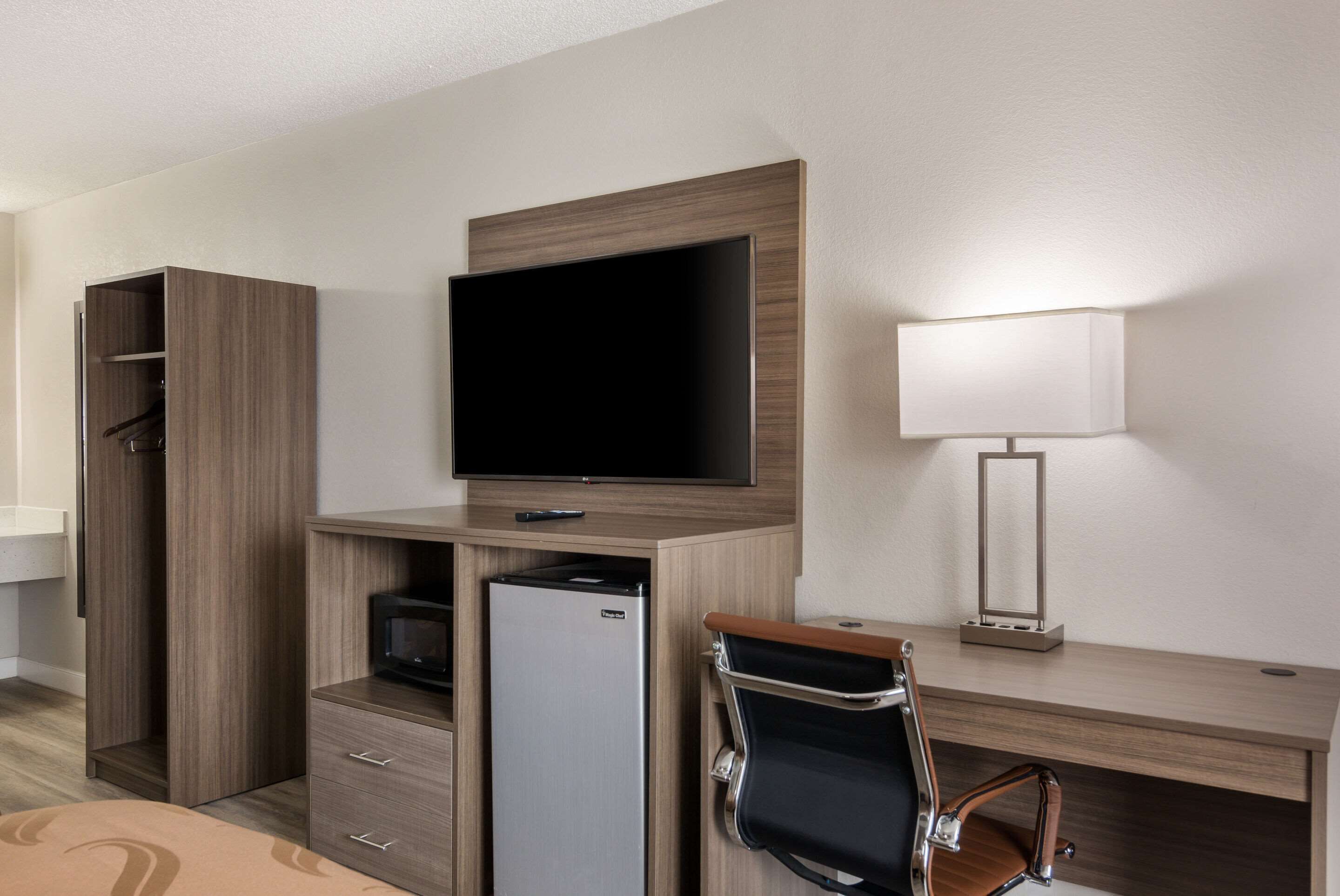 A hotel room with a wooden desk, a flat-screen TV, a refrigerator, a chair, a lamp, and a wardrobe.