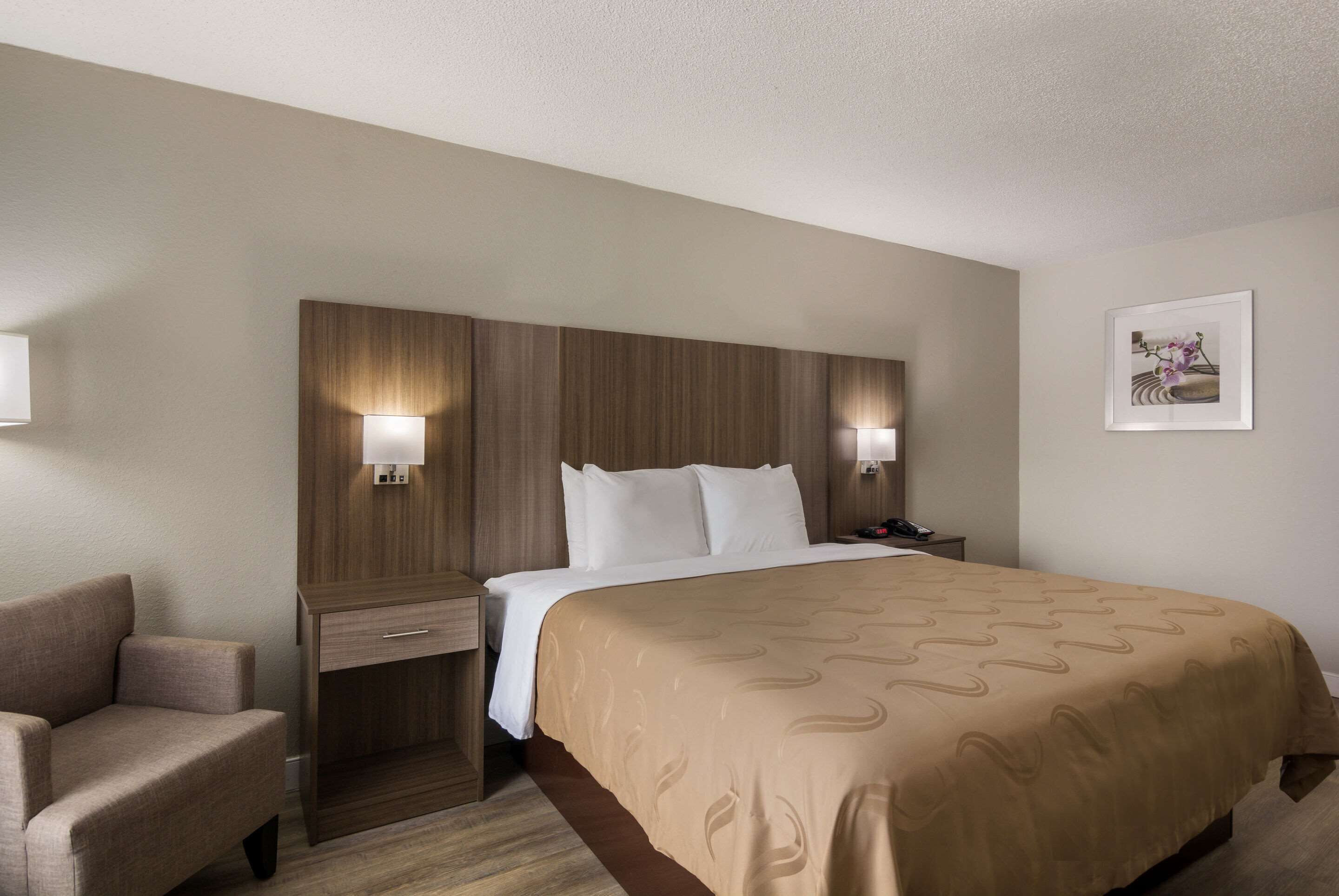 Una habitación de hotel con una cama grande, una mesita de noche, una silla y una lámpara de pared.