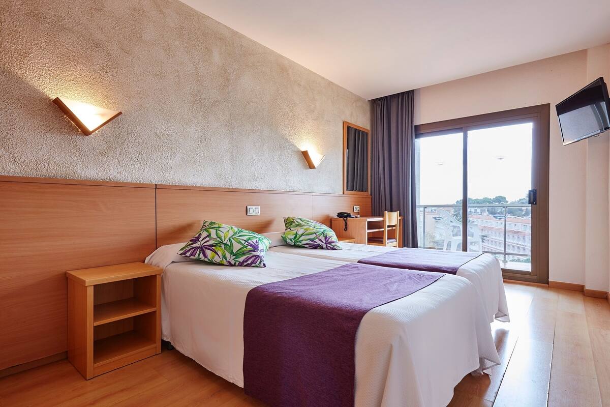 Hotellihuone, jossa on kaksi sänkyä, puinen pääty, yöpöytä, televisio ja suuri ikkuna, josta avautuu näkymä kaupunkiin.