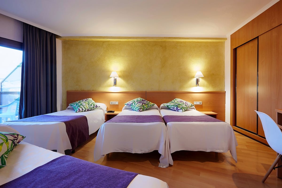 Hotellihuone, jossa on kolme sänkyä, puinen vaatekaappi ja työpöytä tuolilla.