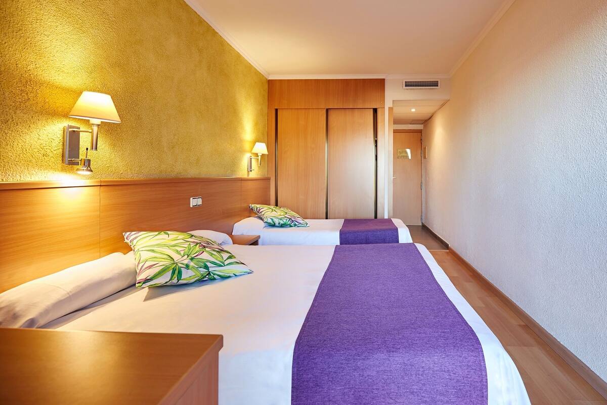 Hotellihuone, jossa on kaksi sänkyä, puinen pääty ja seinään kiinnitetty valaisin sekä puupaneelilla varustettu ovi.