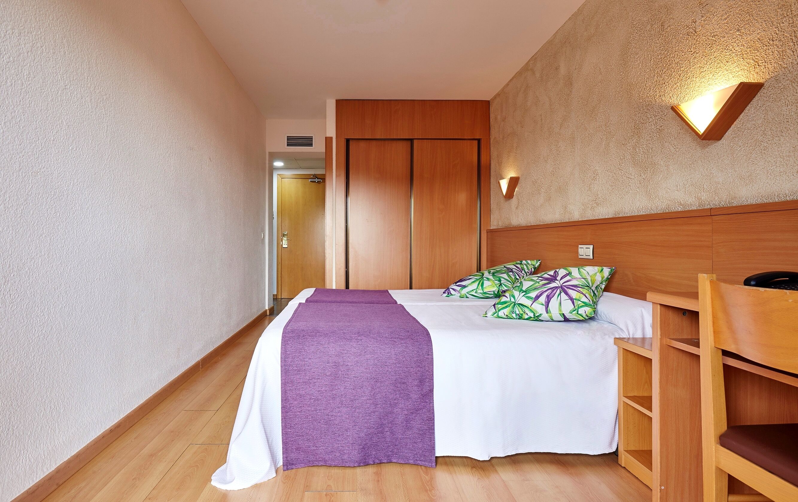 Una habitación de hotel con cama, cabecera de madera, mesita de noche y lámpara fijada a la pared.