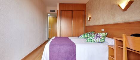 Una habitación de hotel con cama, cabecera de madera, mesita de noche y lámpara fijada a la pared.