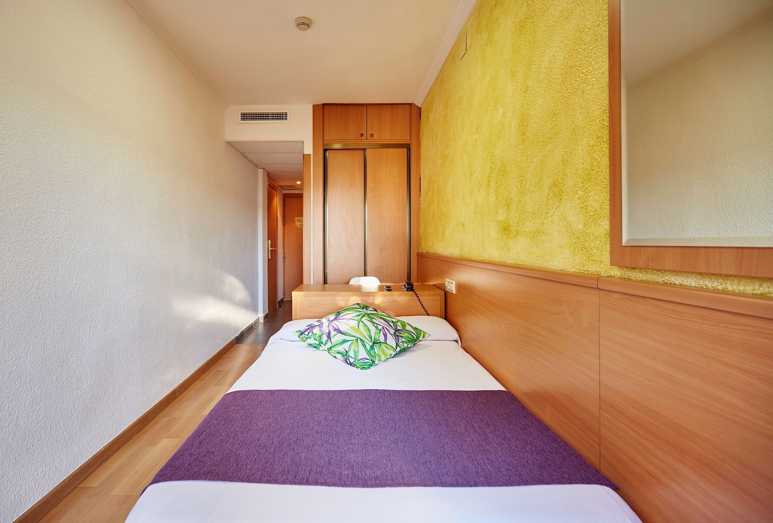 A narrow hallway leading to a bedroom with a bed, a nightstand, and a wooden wardrobe.