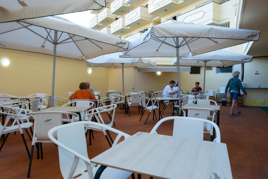 En cafeteria med vita bord och stolar, skyddade av stora, vita parasoller, och en gul vägg i bakgrunden.