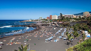 A beach with numerous umbrellas and sun loungers, a rocky shoreline, and buildings in the background.