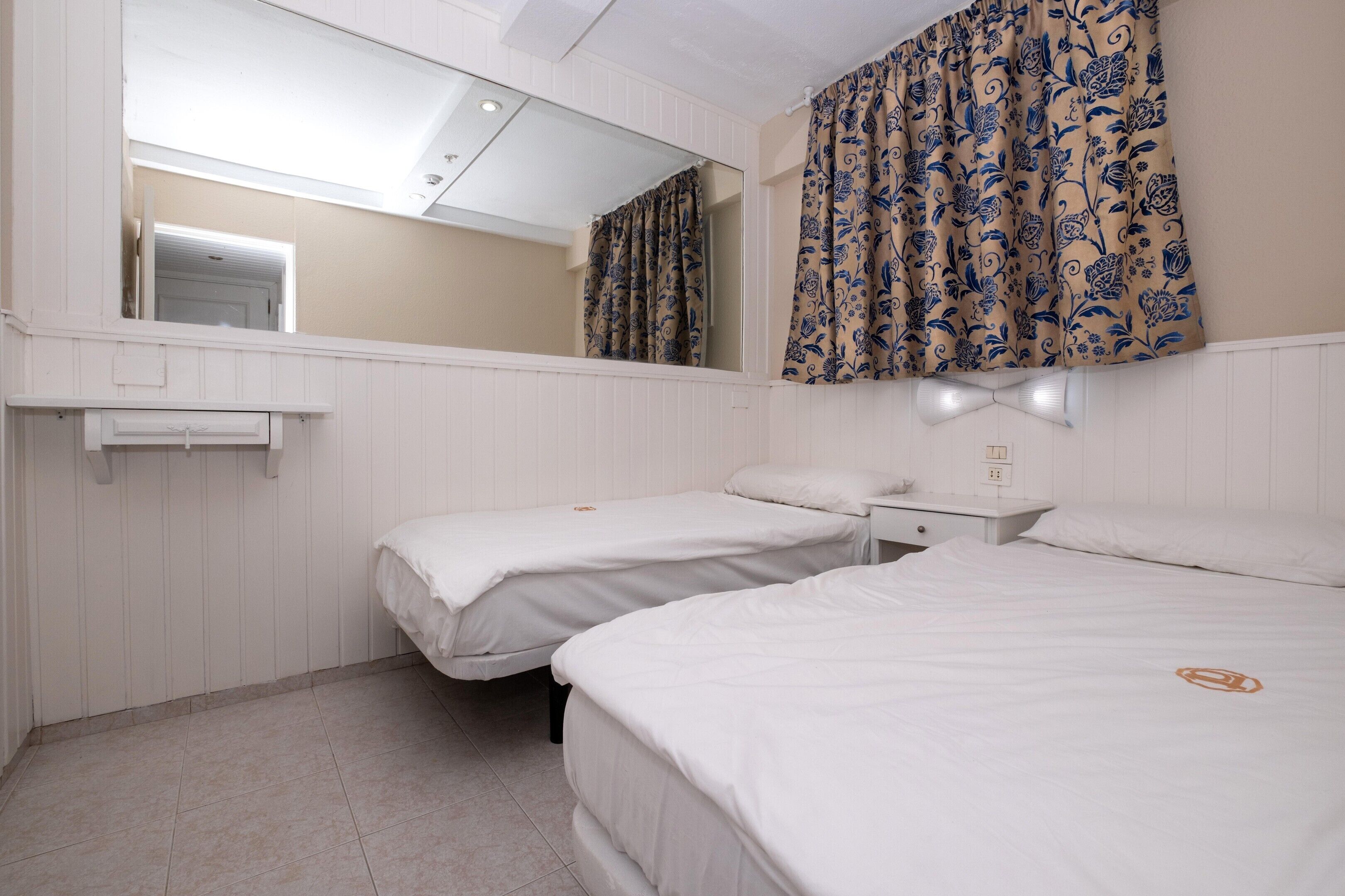 Habitación de hotel con dos camas, un espejo, una mesita de noche, una lámpara y una ventana con cortinas.