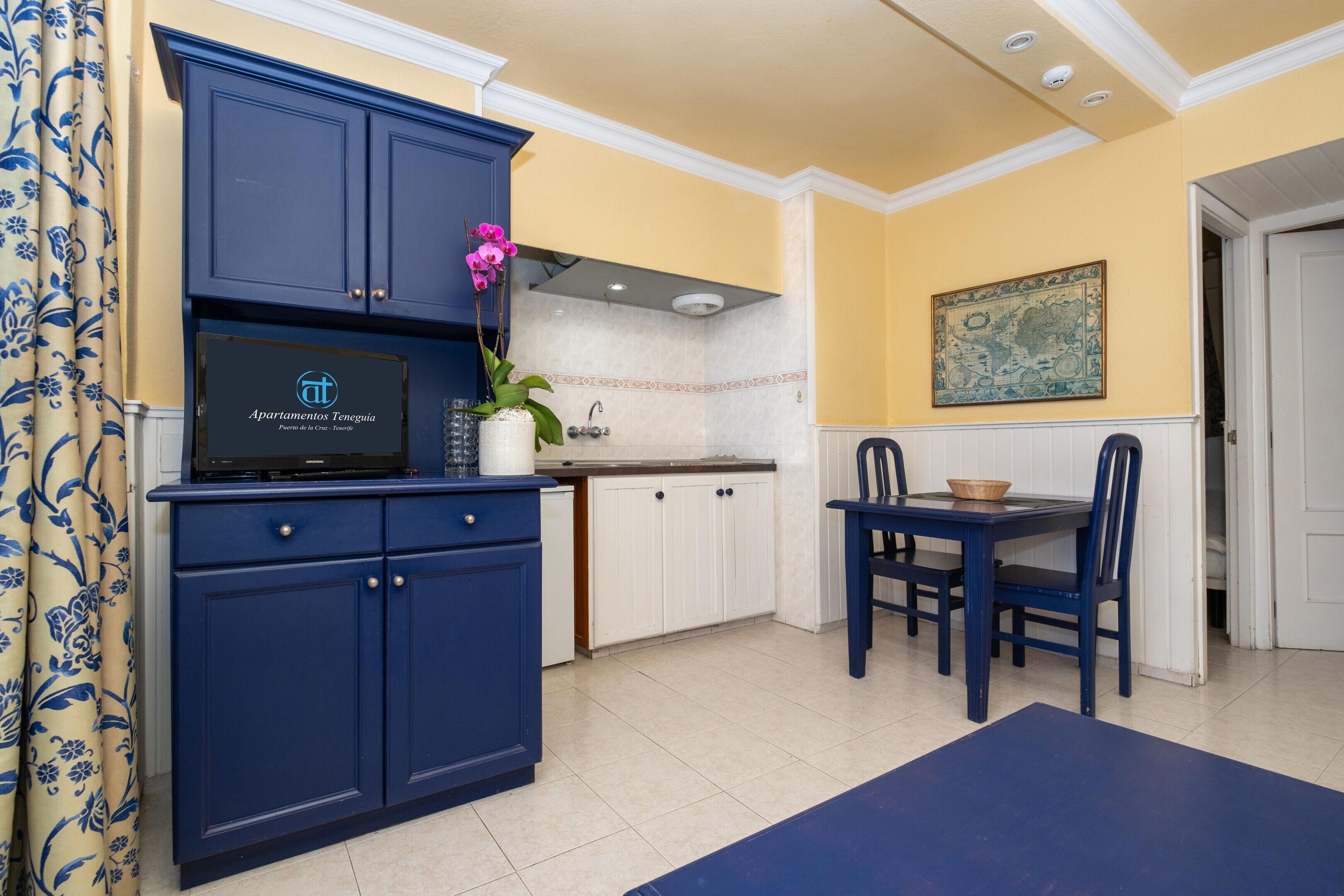 Una cocina con armarios azules, una mesa de comedor con sillas, una planta en maceta y un adorno en la pared.