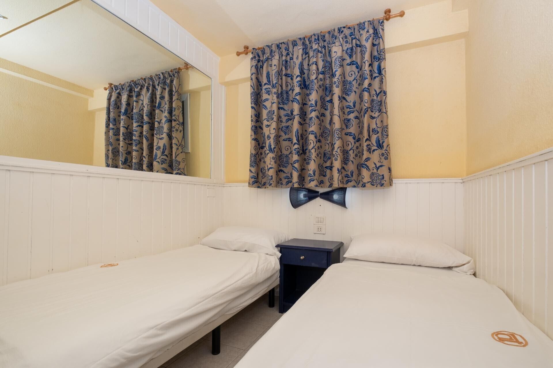 Une chambre avec deux lits simples, une table de chevet, un miroir et des rideaux à motifs.