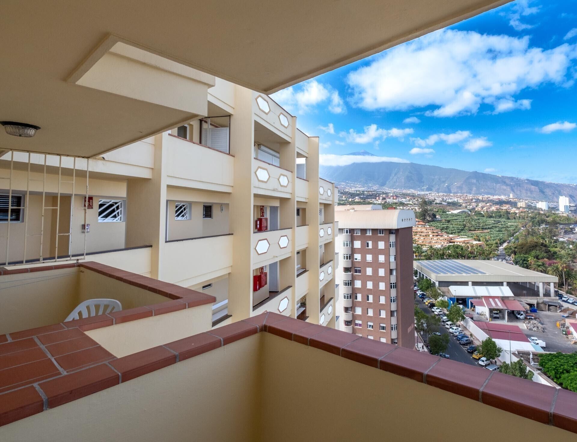 A multi-story building with balconies, a view of a mountainous landscape, and a parking area with vehicles.