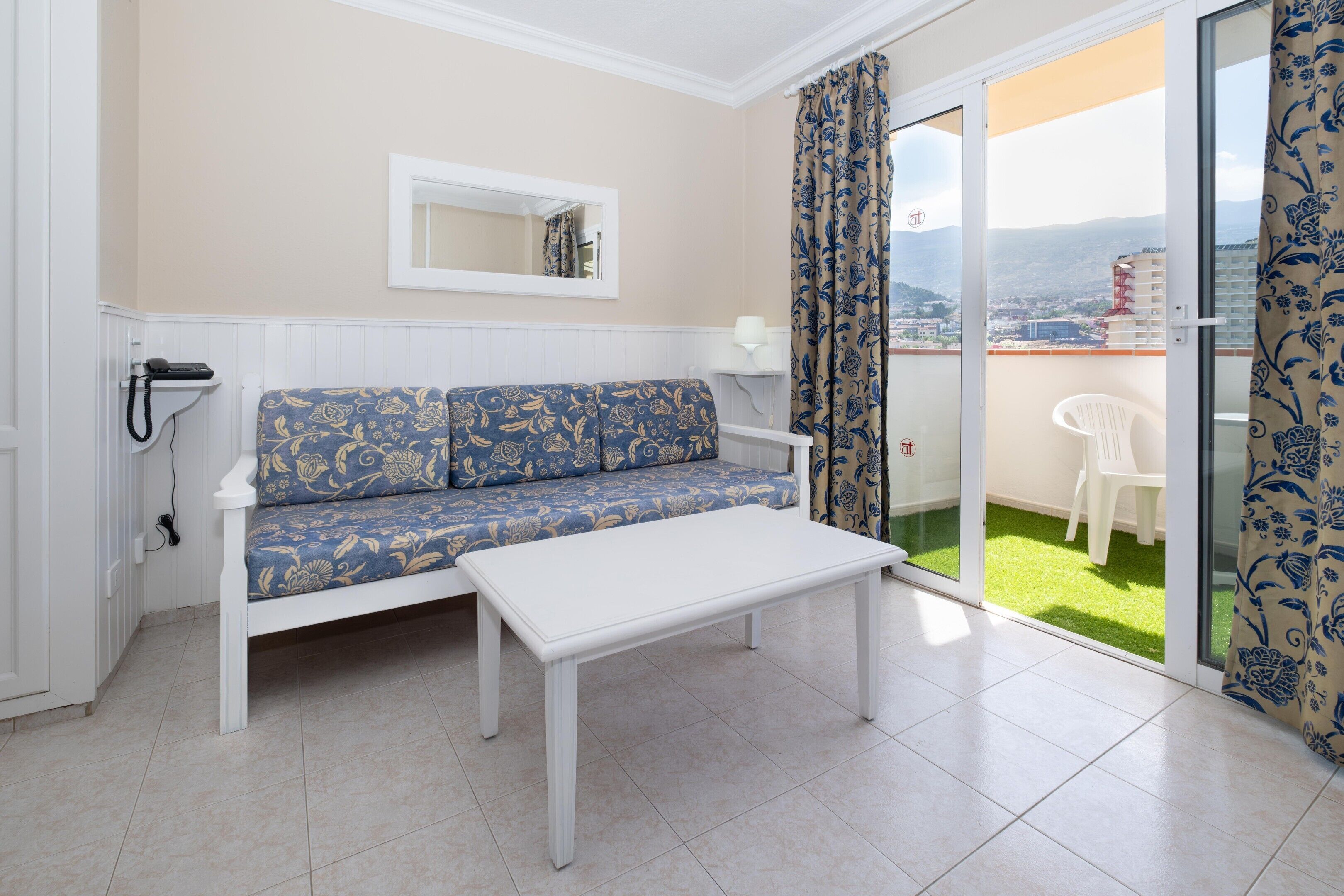 A room with a sofa, a table, a mirror, and a balcony with a view.