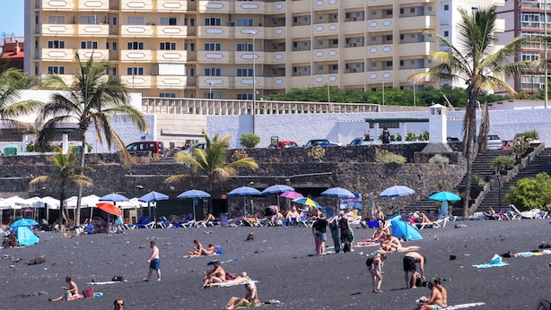 A multi-story hotel with balconies, a beach in the foreground, and palm trees.