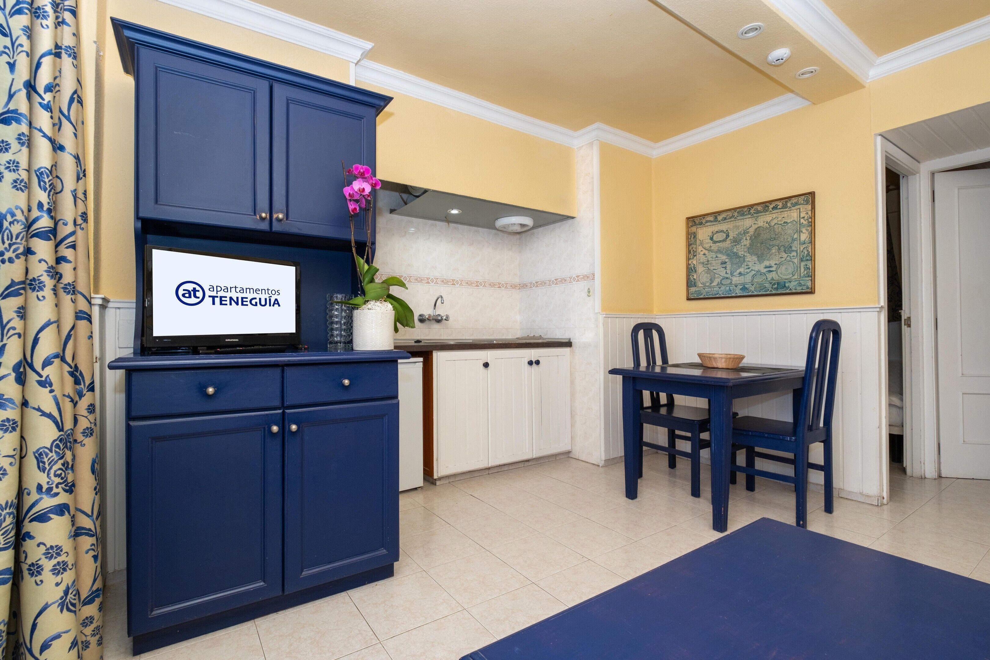 Une cuisine avec des meubles bleus, une table à manger avec des chaises, une orchidée en pot et une enseigne indiquant « apartamentos TENERGUIA ».