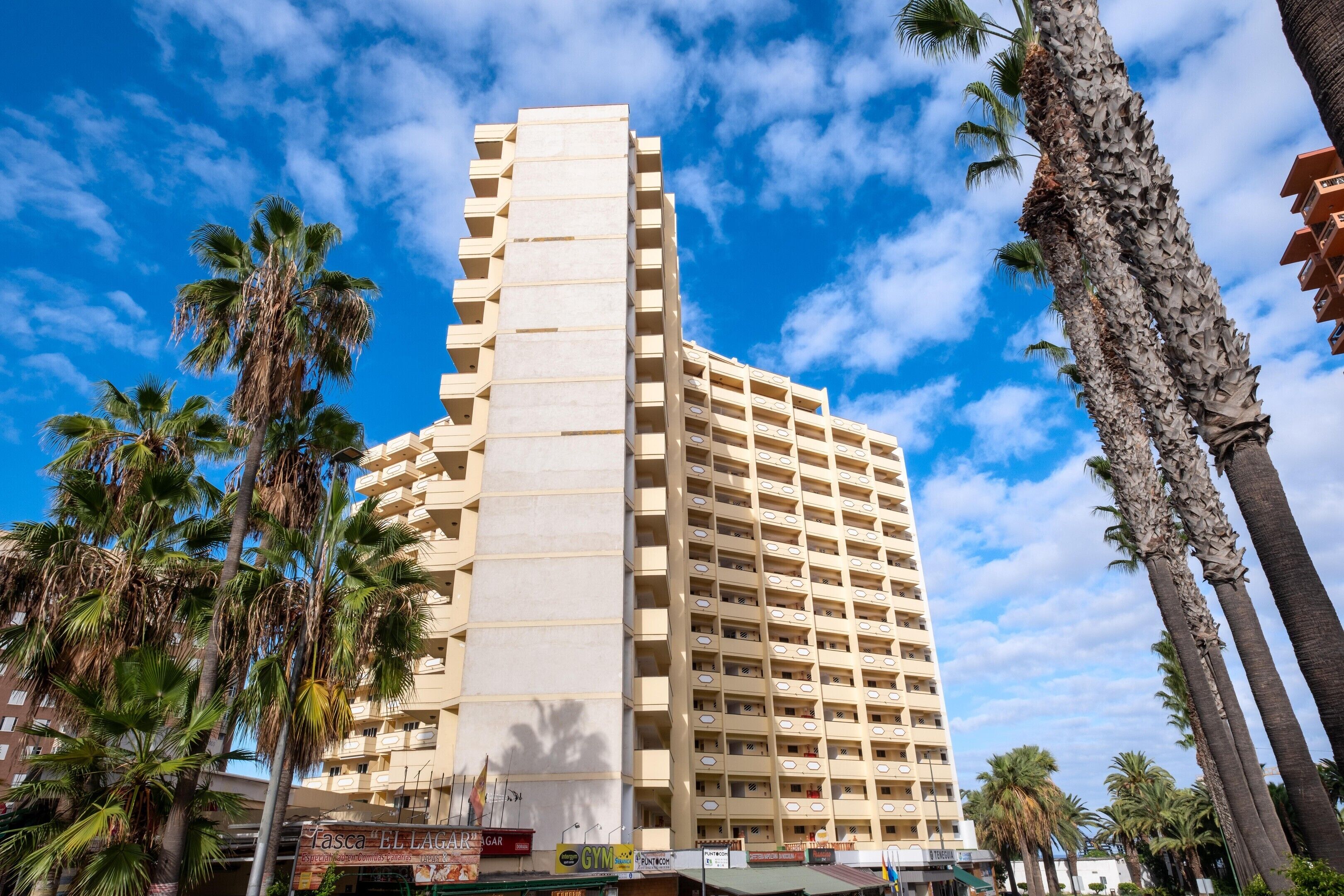 A tall, beige multi-story building with balconies, surrounded by palm trees.