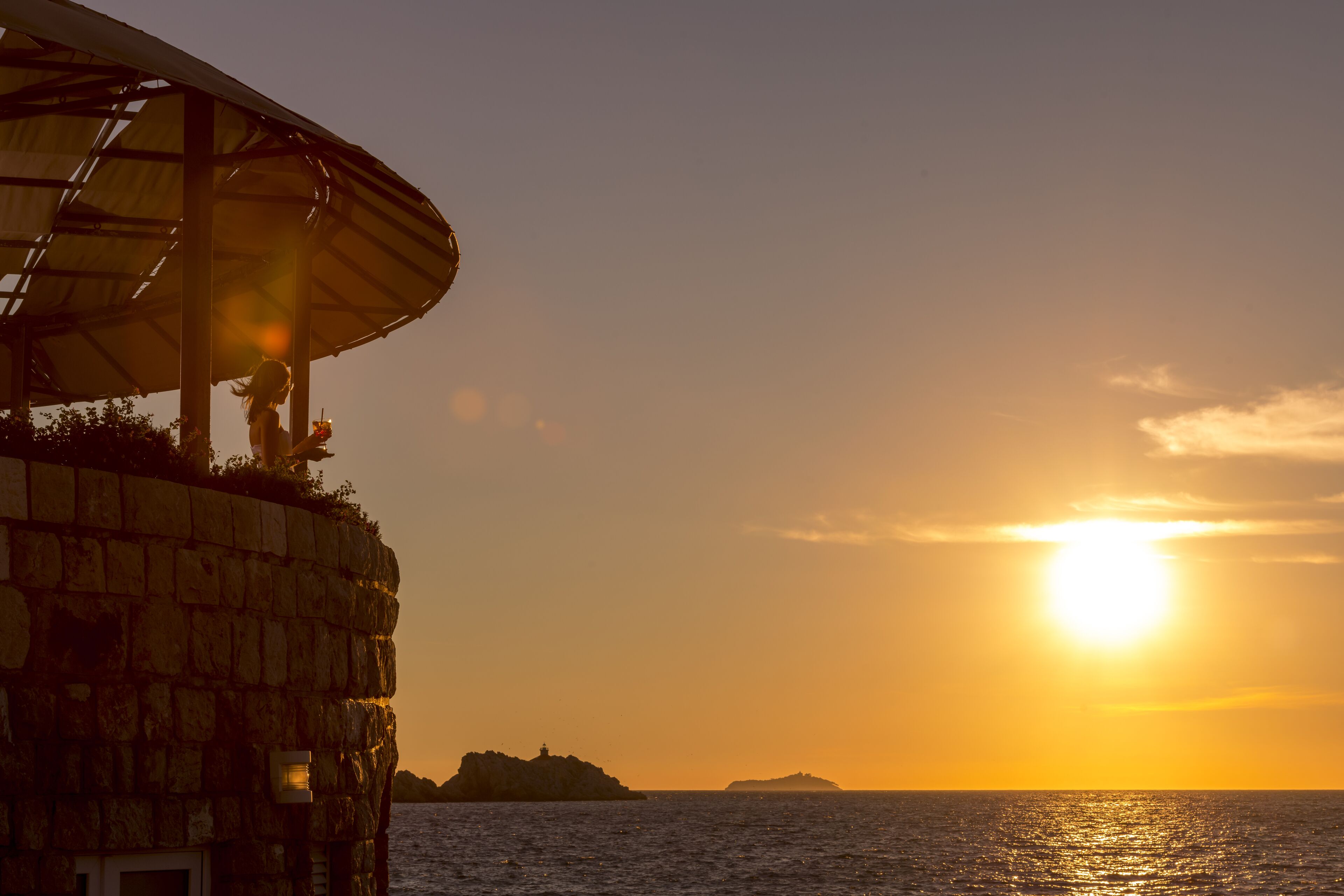 Ein Steinhaus mit einer großen Sonnenschirmkonstruktion, mit Blick auf ein Gewässer im Sonnenuntergang.