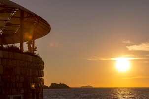 Um edifício de pedra com uma grande estrutura de guarda-sol, com vista para um corpo de água e um pôr do sol ao fundo.