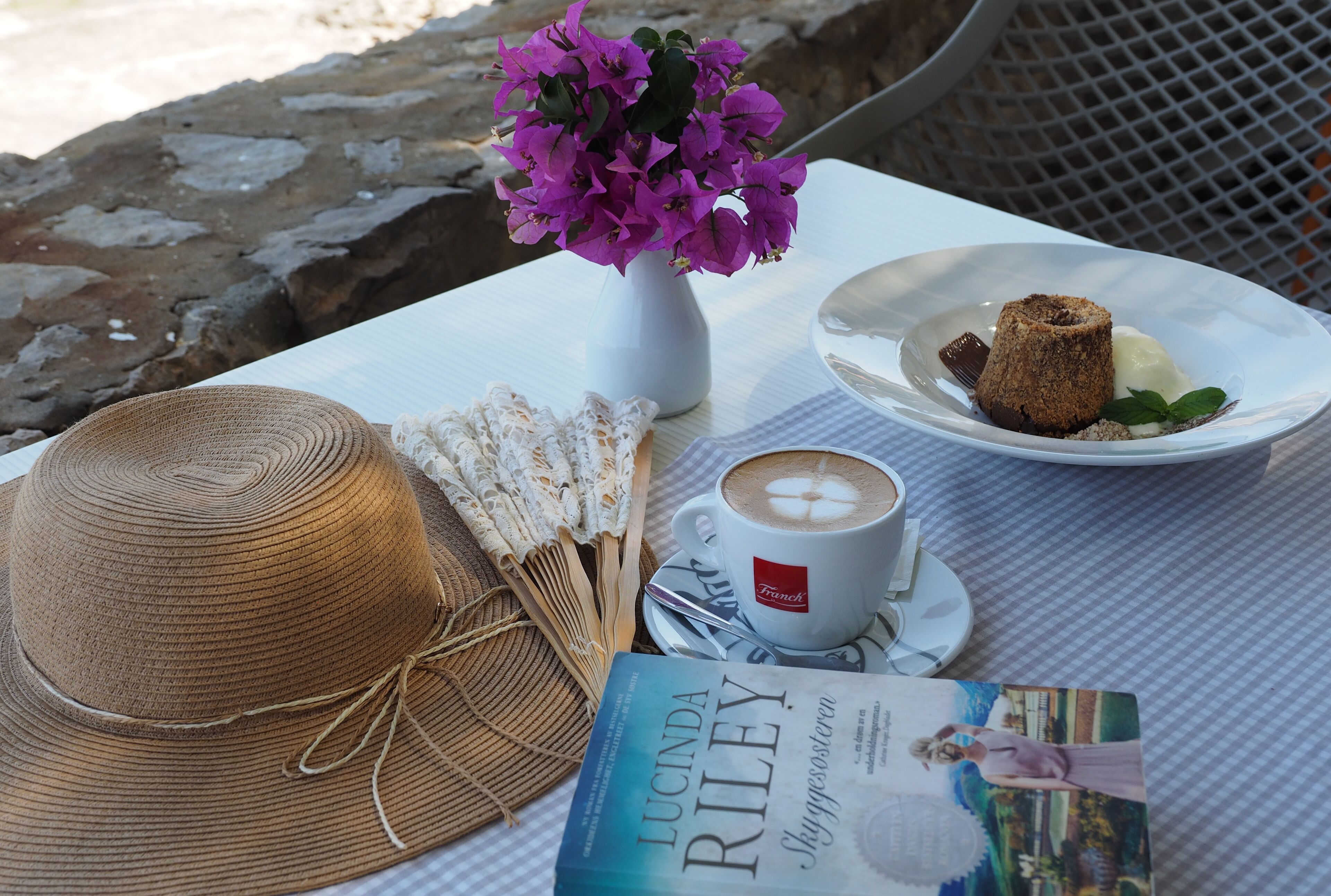 A table with a book, a hat, a cup of coffee, a plate with a dessert, and a vase with flowers.