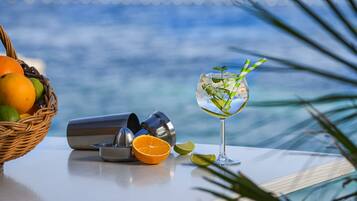 A table with a cocktail glass, a cocktail shaker, and a basket of fruits by a window overlooking the ocean.